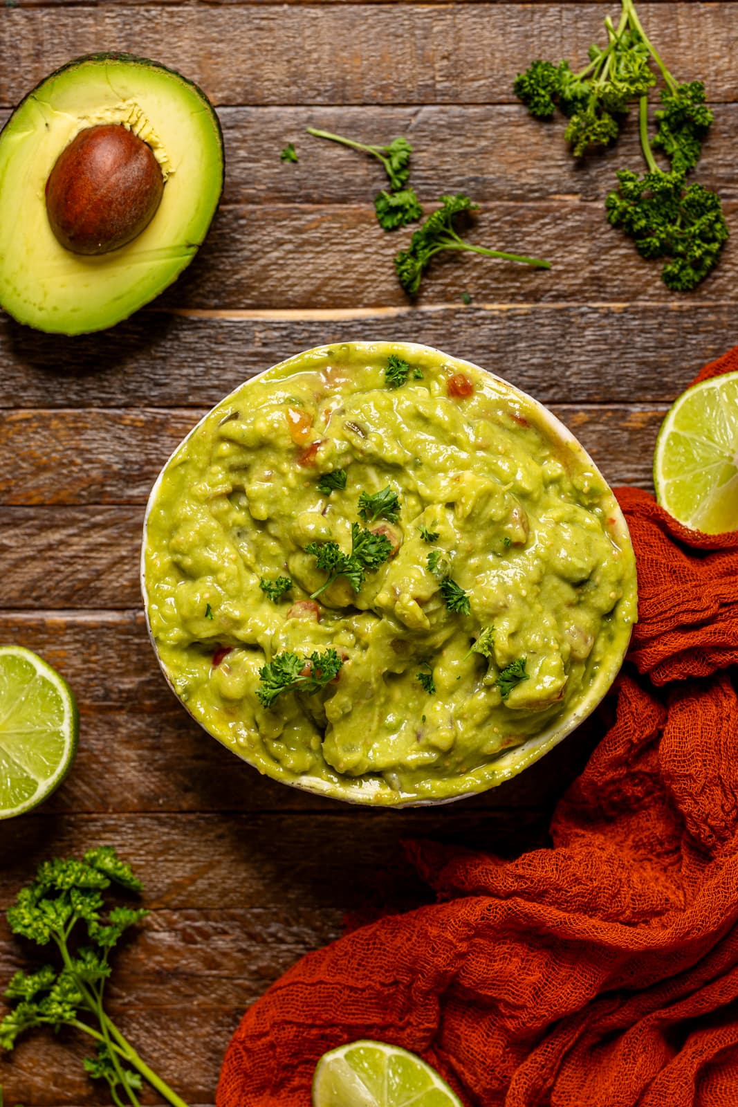 A bowl of guacamole on a brown wood table with limes and avocado.