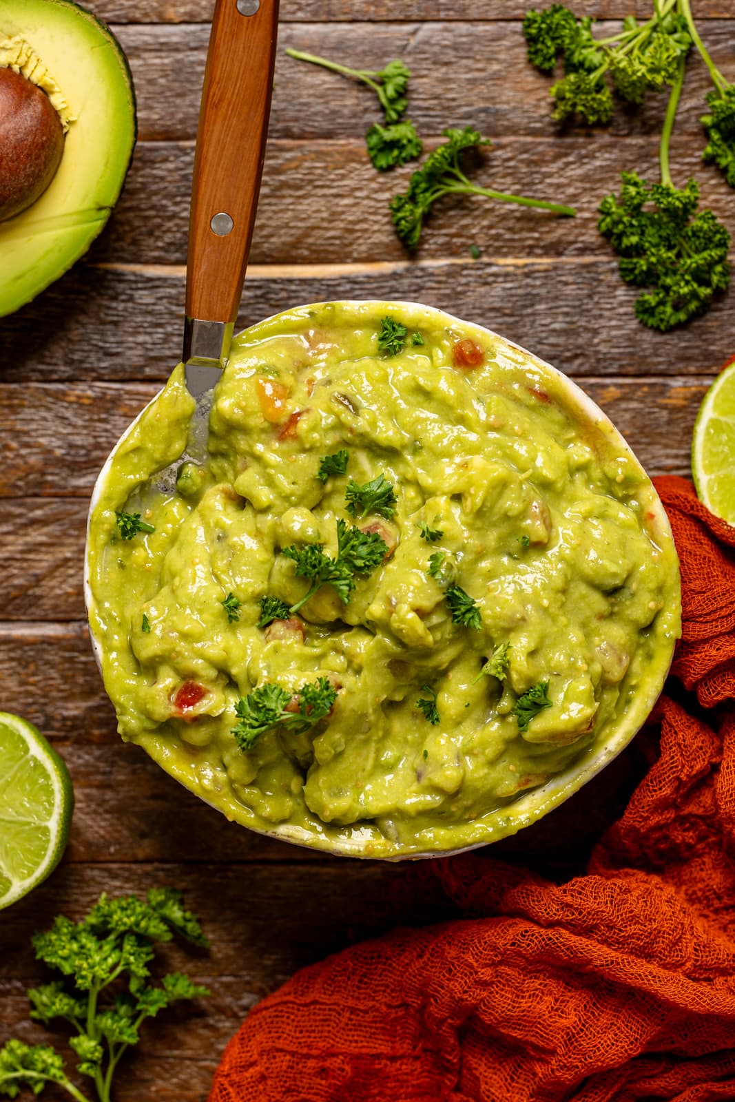 Guacamole in a bowl with a spoon with lime, fresh herbs, and avocado.