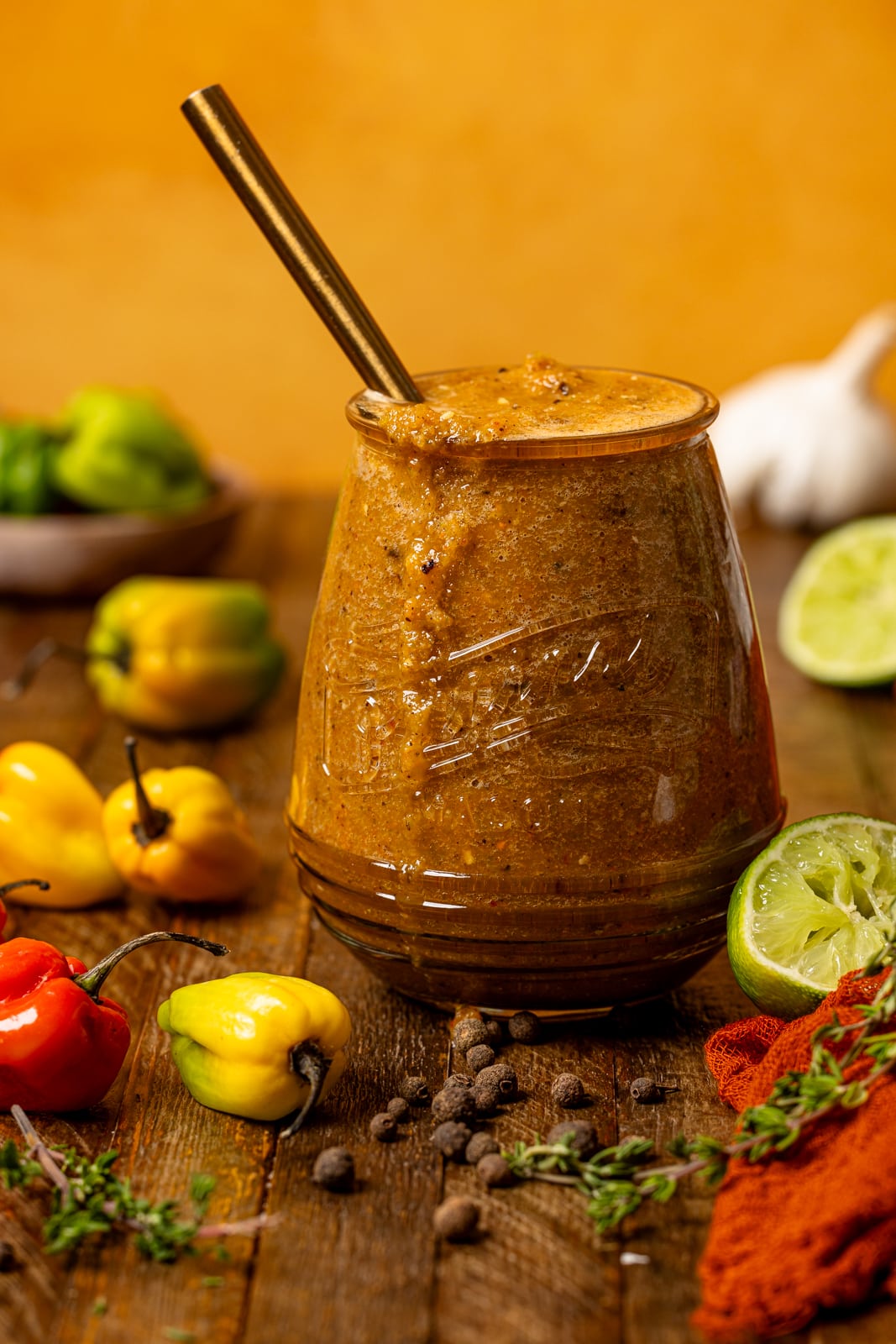 Pepper sauce in a mason jar with a spoon and scotch bonnet and thyme sprigs on a brown wood table.