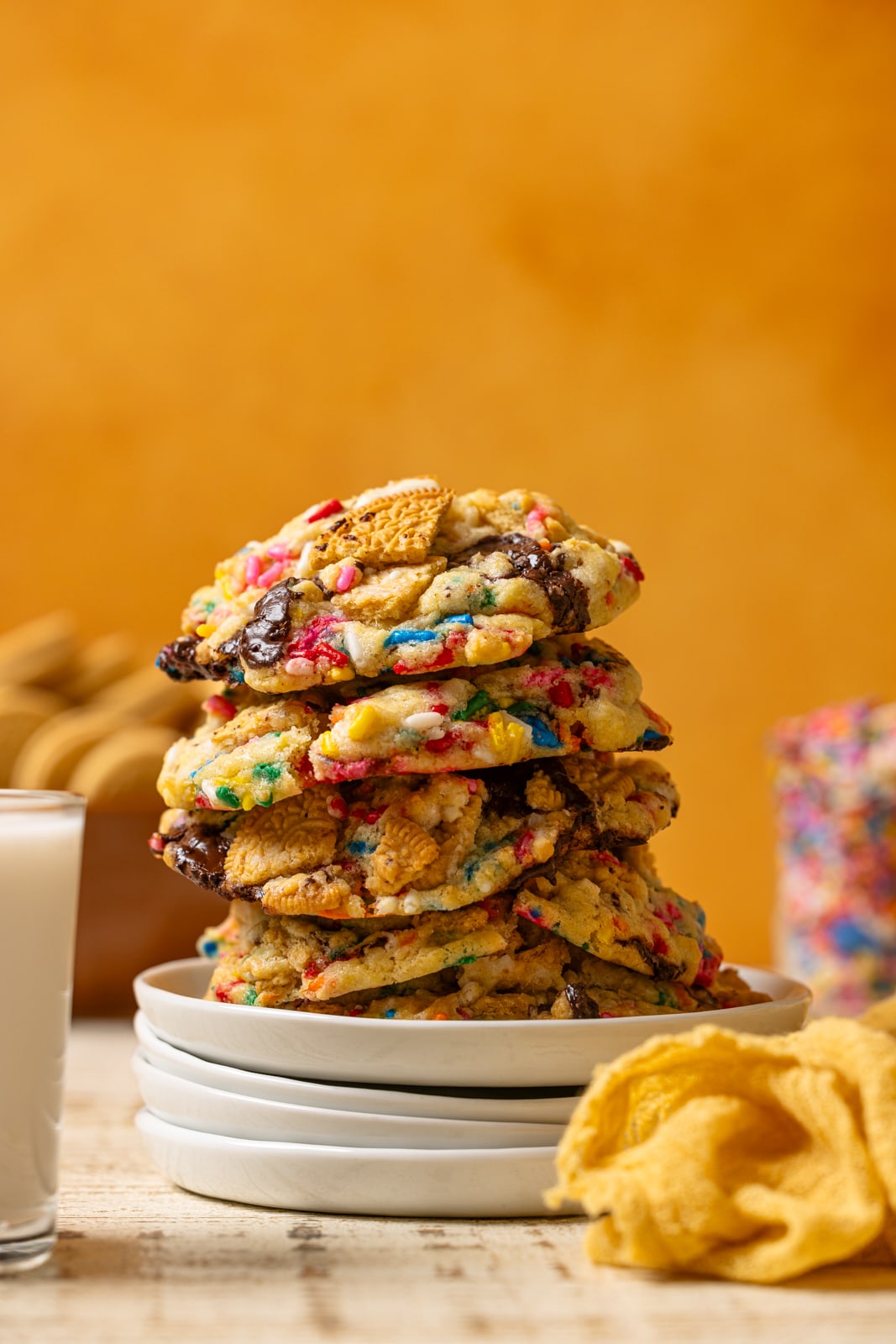 Stacked cookies on a white plates with a glass of milk and yellow napkin.