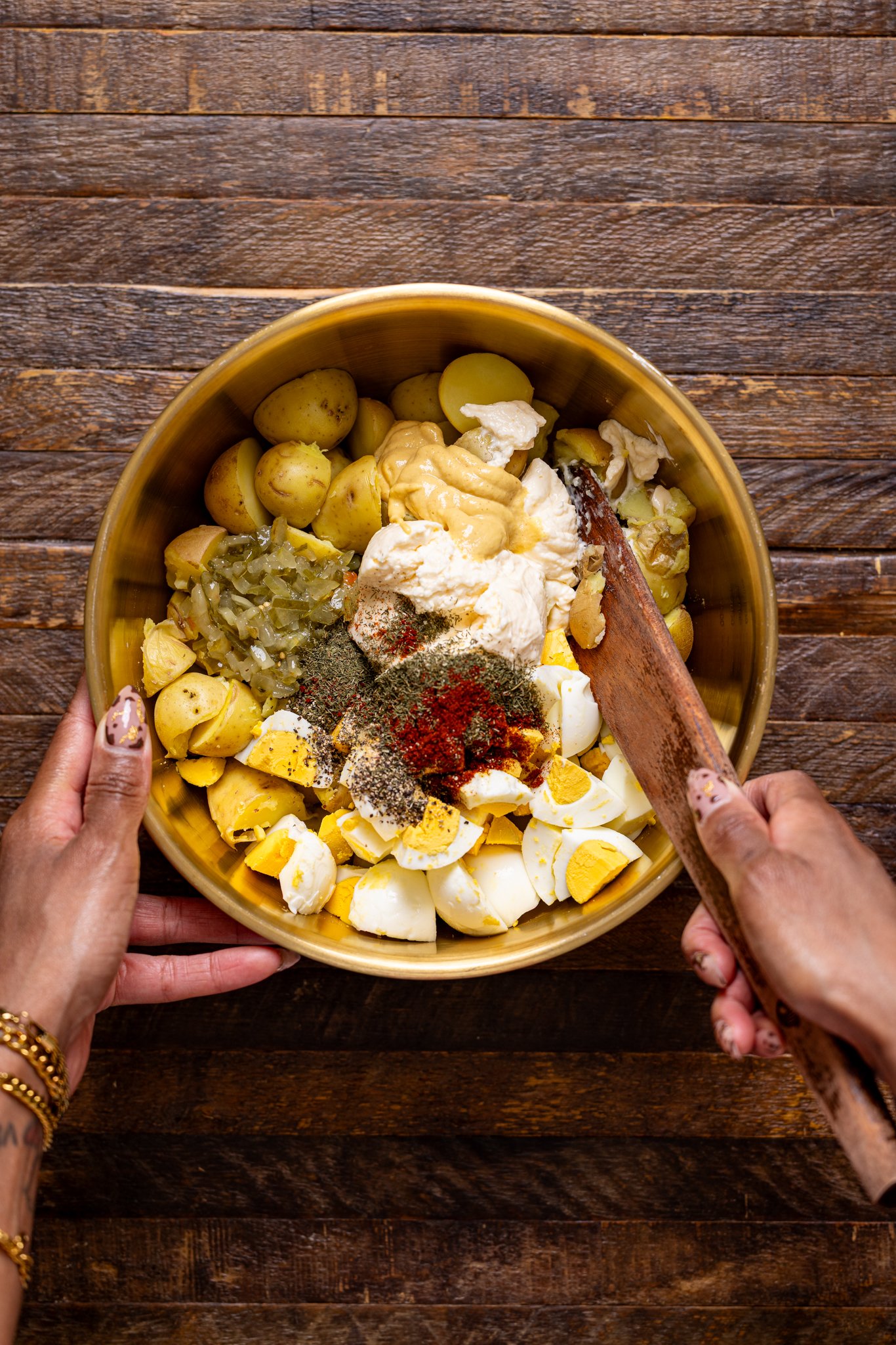 Potato salad ingredients together in a bowl with a spoon being held by woman hands.