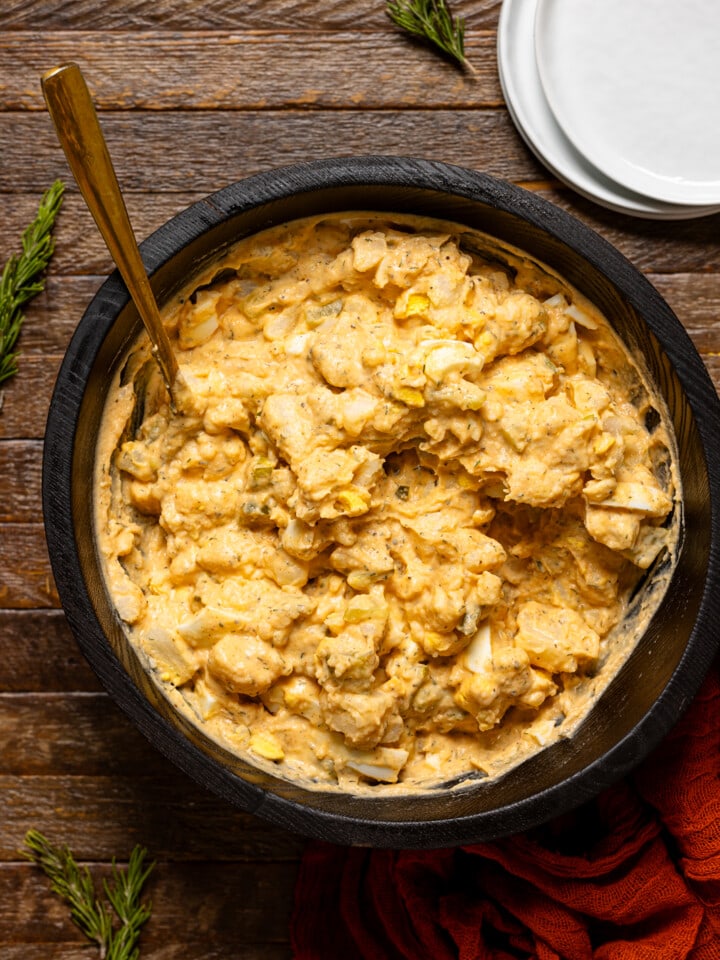 Potato salad in a black bowl with a gold spoon and white plates on the side.