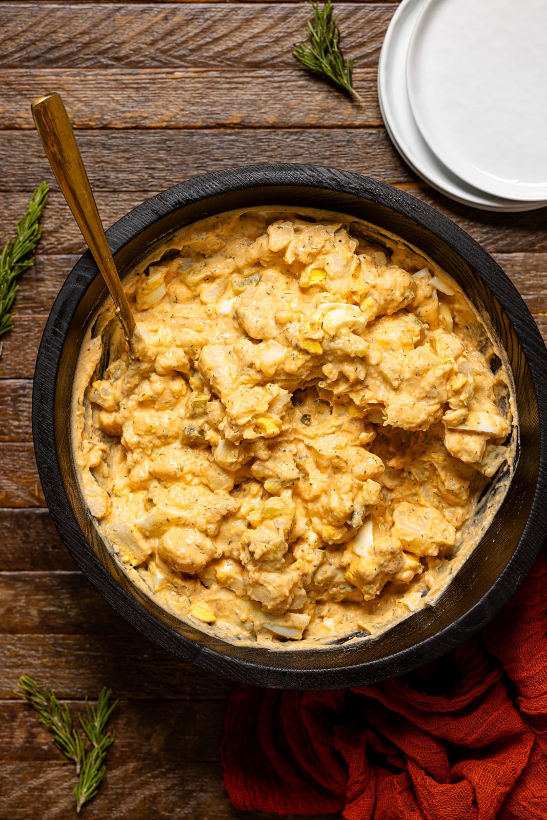 Potato salad in a black bowl with a gold spoon and white plates on the side.