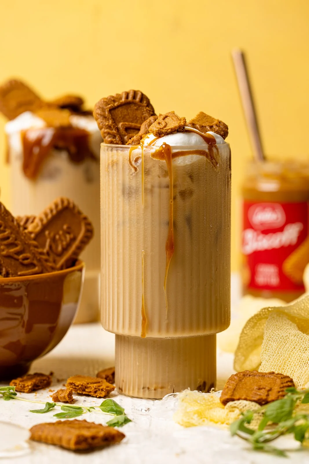 Two glasses with lattes, biscoff cookies, and cookie butter in the background on a white table and yellow background.