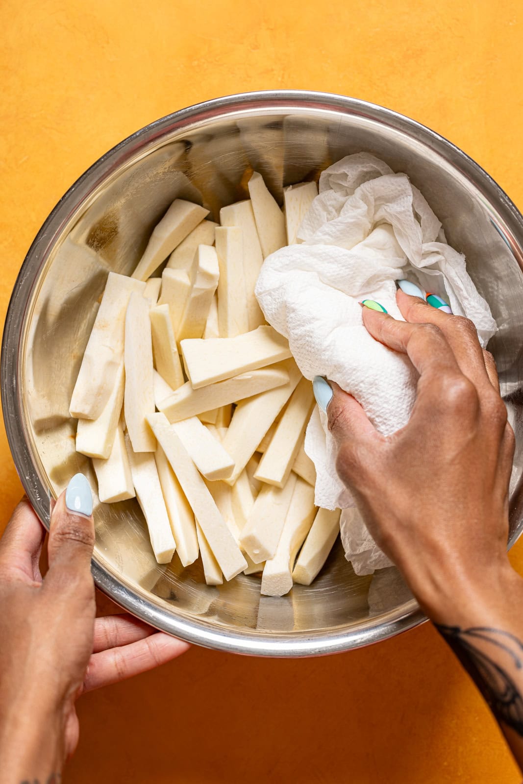 Sliced Yucca being patted dry with a paper towel in a silver bowl.