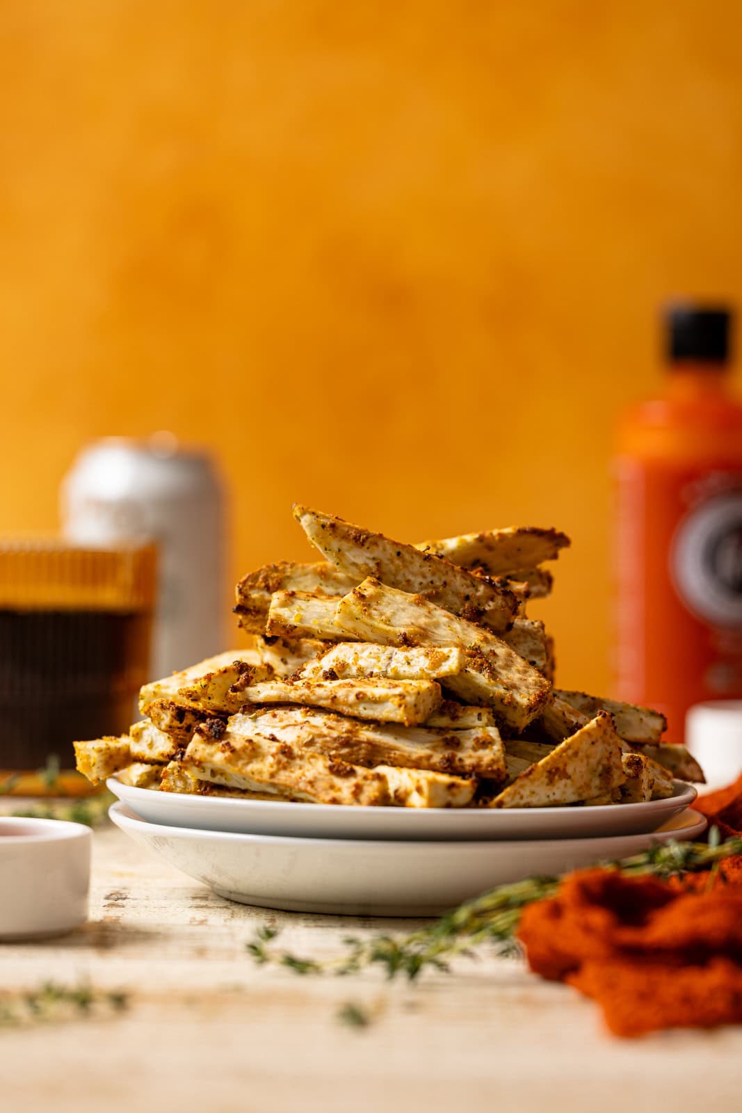 Yucca fries stacked atop white plates with an orange background, drink, and hot sauce in the background. 