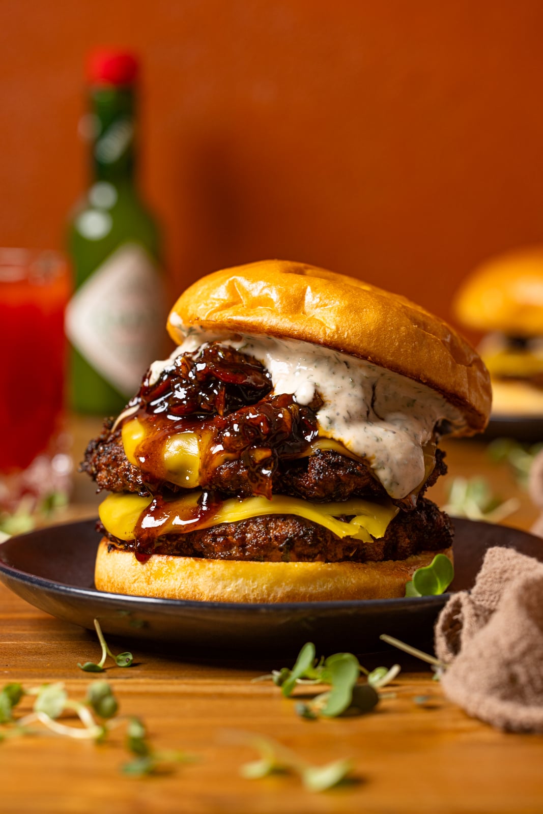 Burger on a plate on a brown wood table with a drink, tobasco sauce, and burger in the background.