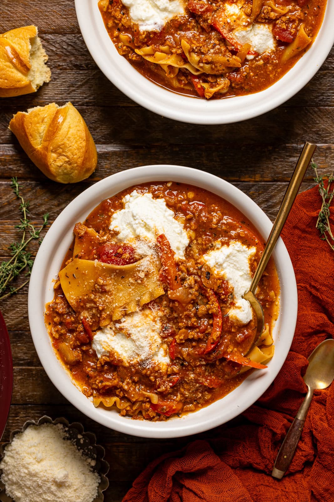 Two bowls of lasagna soup with a spoon and bread on a brown wood table.