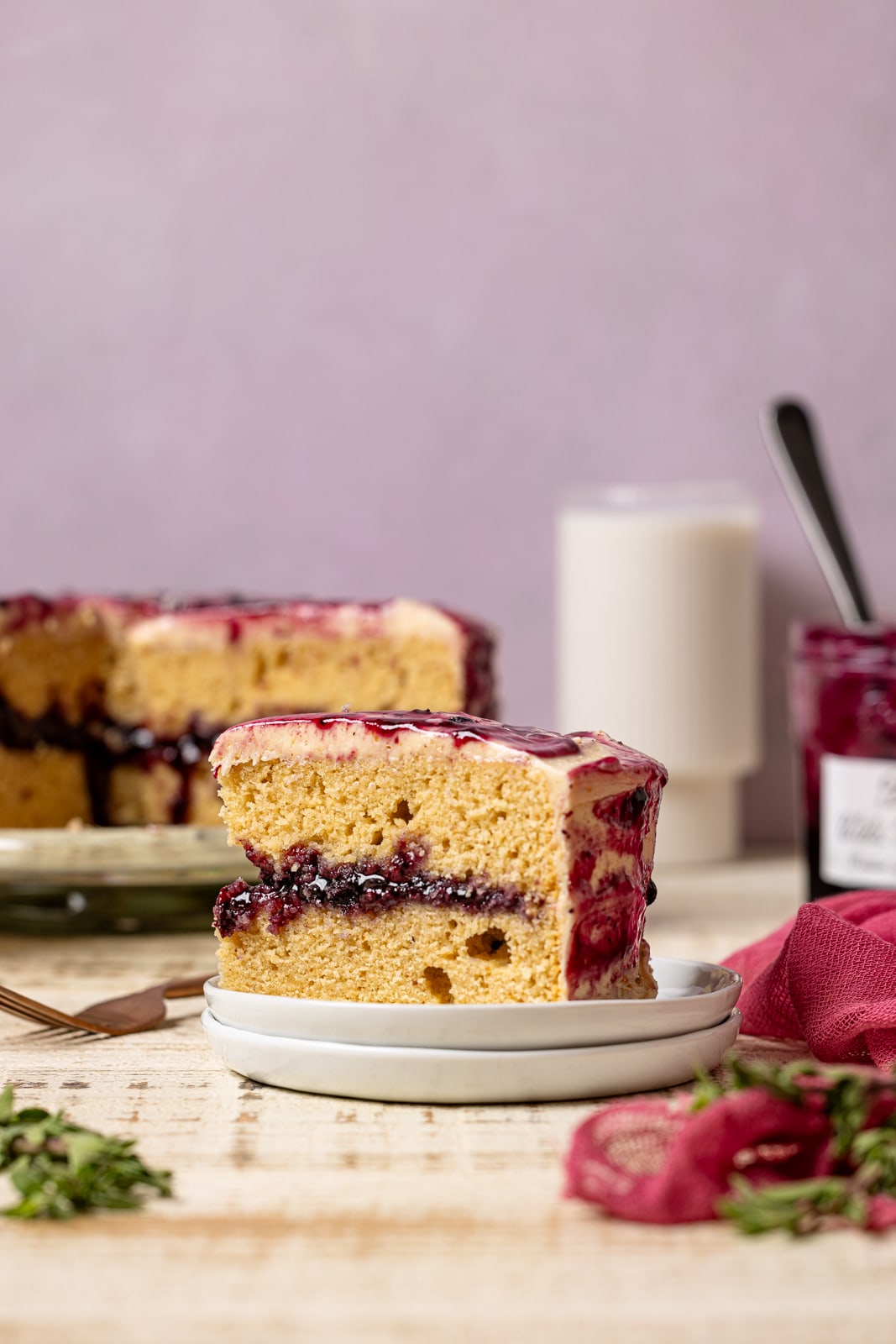 A cake slice on a stack of white plates with cake, jam, and milk in the background. 