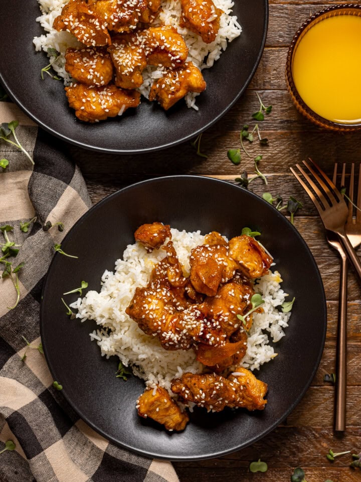 Two plates of Orange chicken with forks and a glass of juice.