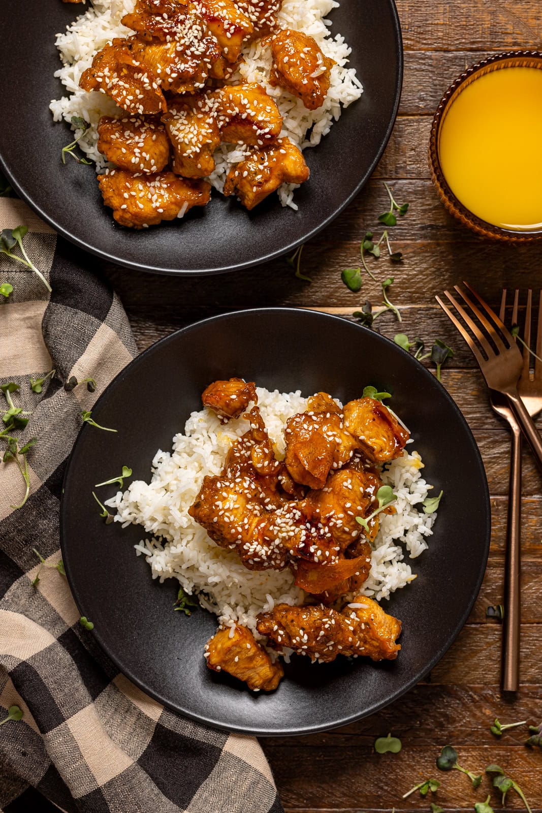 Two plates of Orange chicken with forks and a glass of juice.