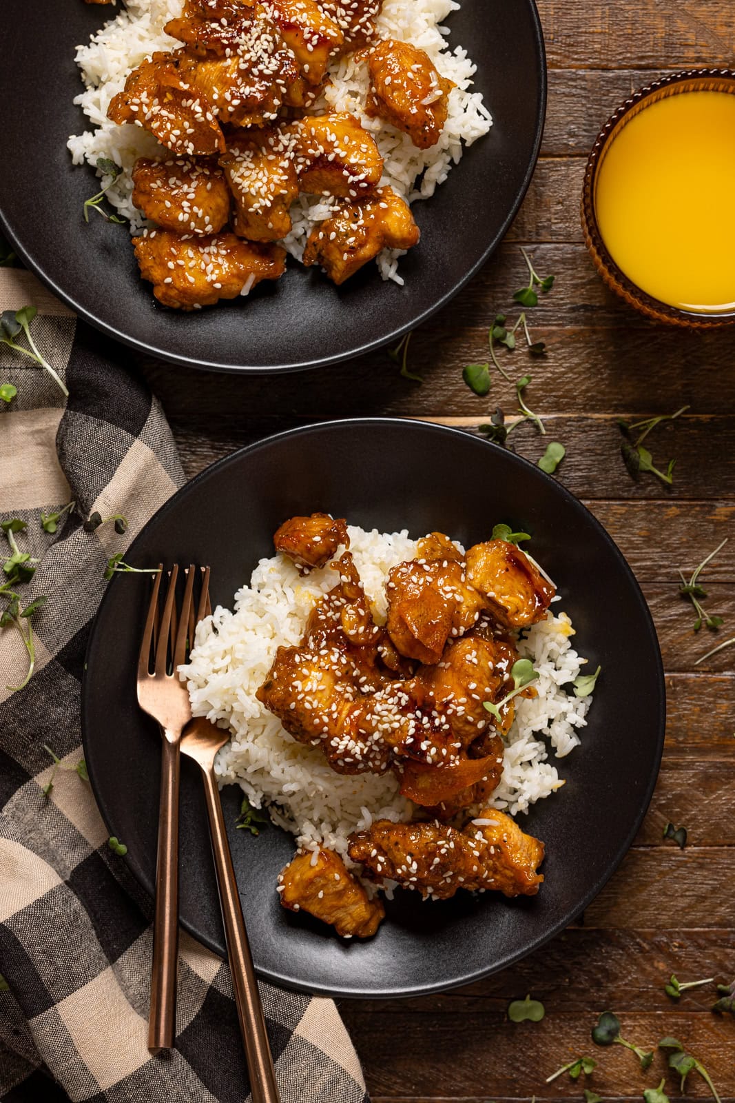 Two plates of Orange Chicken with forks and juice on a brown wood table.