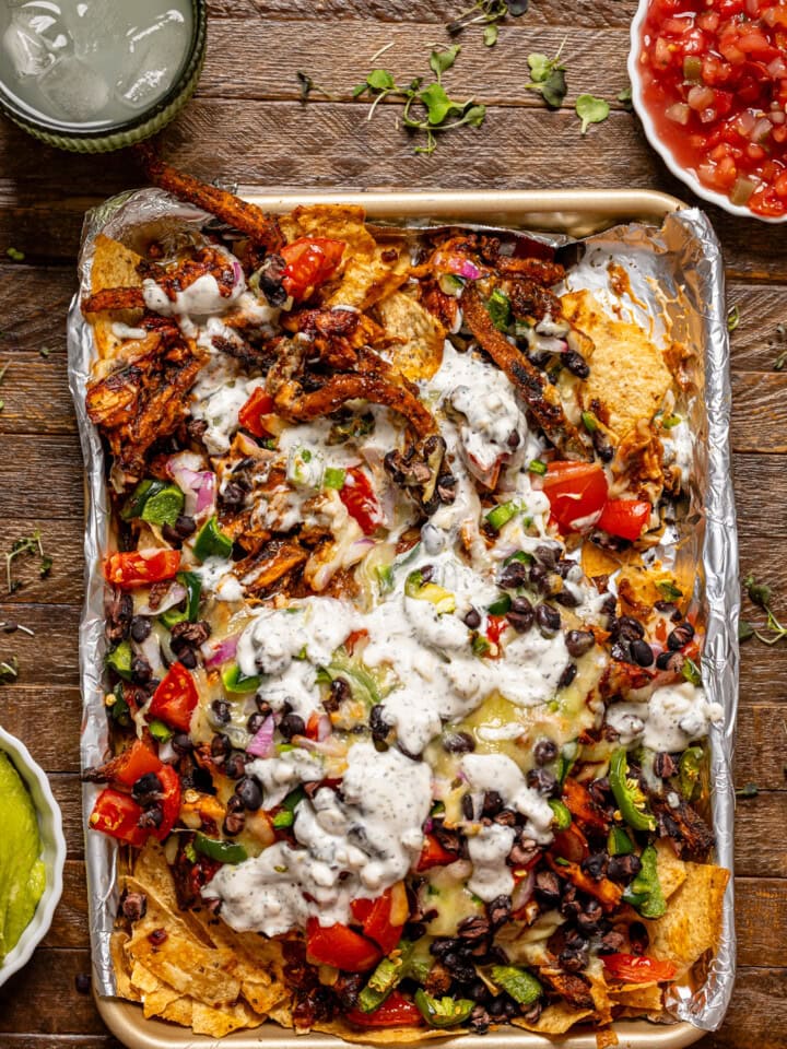 Nachos on a baking sheet with a drink and a side of guacamole and salsa on a brown wood table.