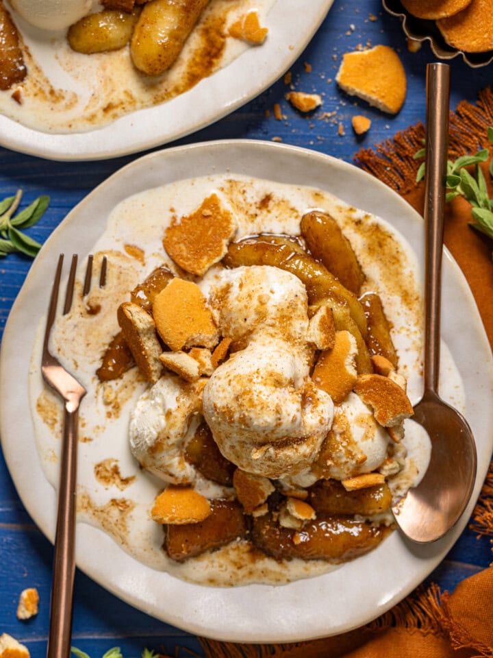 Bananas foster in a white plates with a fork and spoon and cookies on a blue wood table.