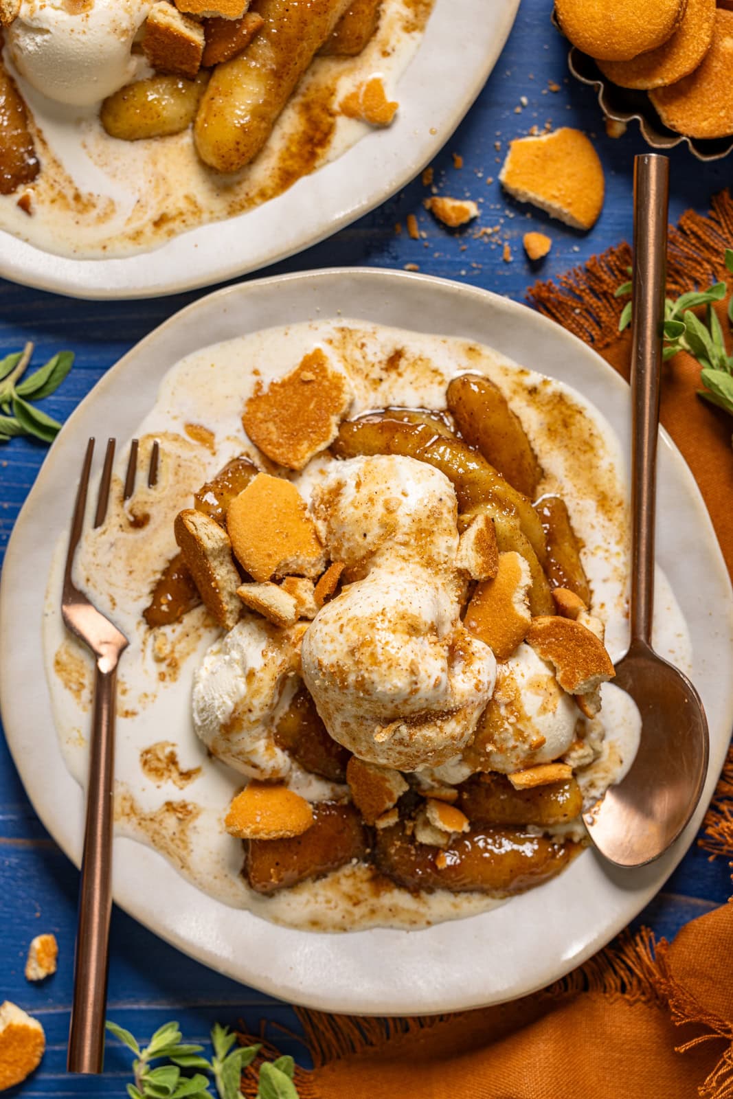Bananas foster in a white plates with a fork and spoon and cookies on a blue wood table.