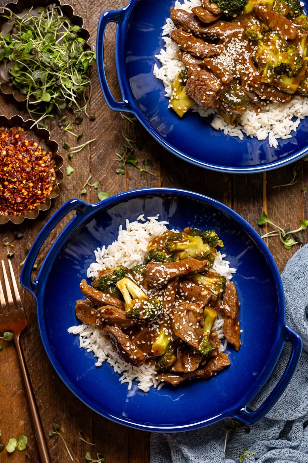 Two blue plates on beef and broccoli on a brown wood table with a fork.