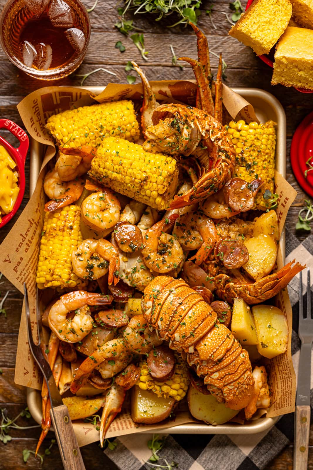Up close shot of seafood boil on a baking sheet with a drink, cornbread, and mac + cheese on a brown wood table.