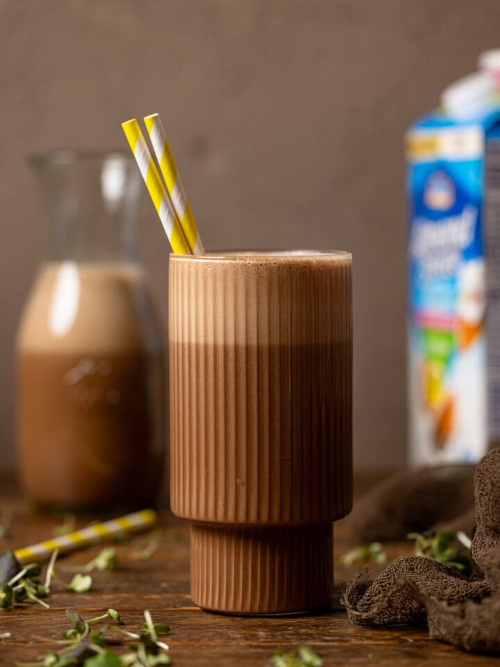 Chocolate milk in a glass with a yellow stripe straw and in a jar in the background with a carton of milk.