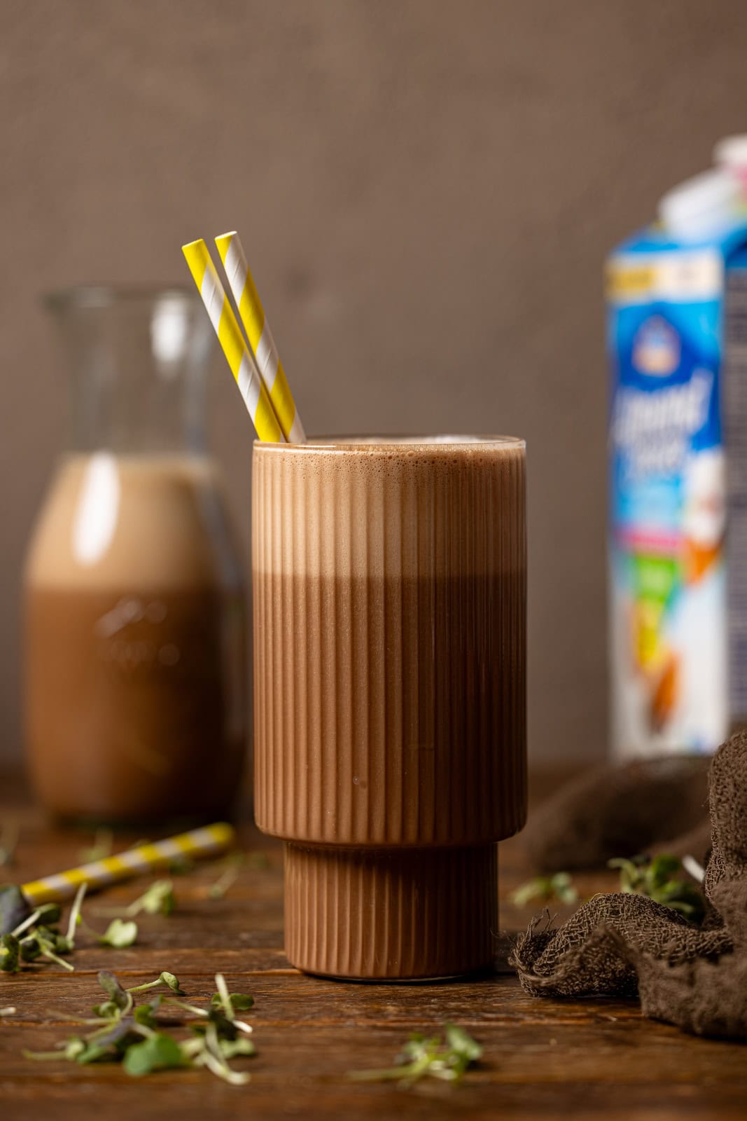 Chocolate milk in a glass with a yellow stripe straw and in a jar in the background with a carton of milk.