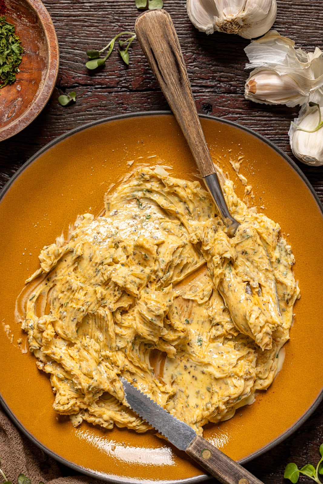 Herb butter on a plate on a dark brown wood table with garlic and seasonings.