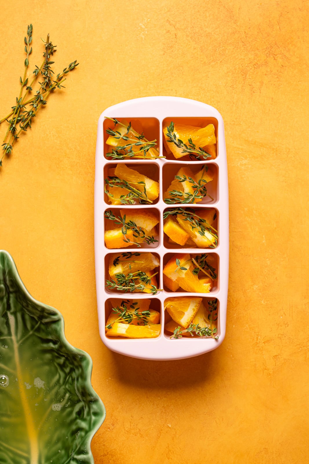 Oranges, thyme, and water in an ice tray on an orange table.