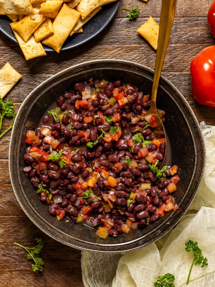 Black bean salad in a bowl with a gold spoon, crackers, and tomatoes.