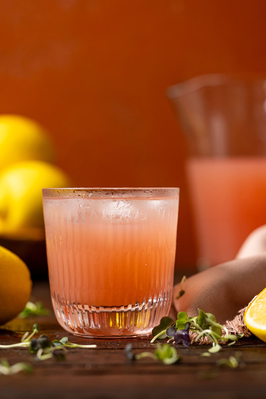 Glass and pitcher of pink lemonade with lemons and herb garnish.