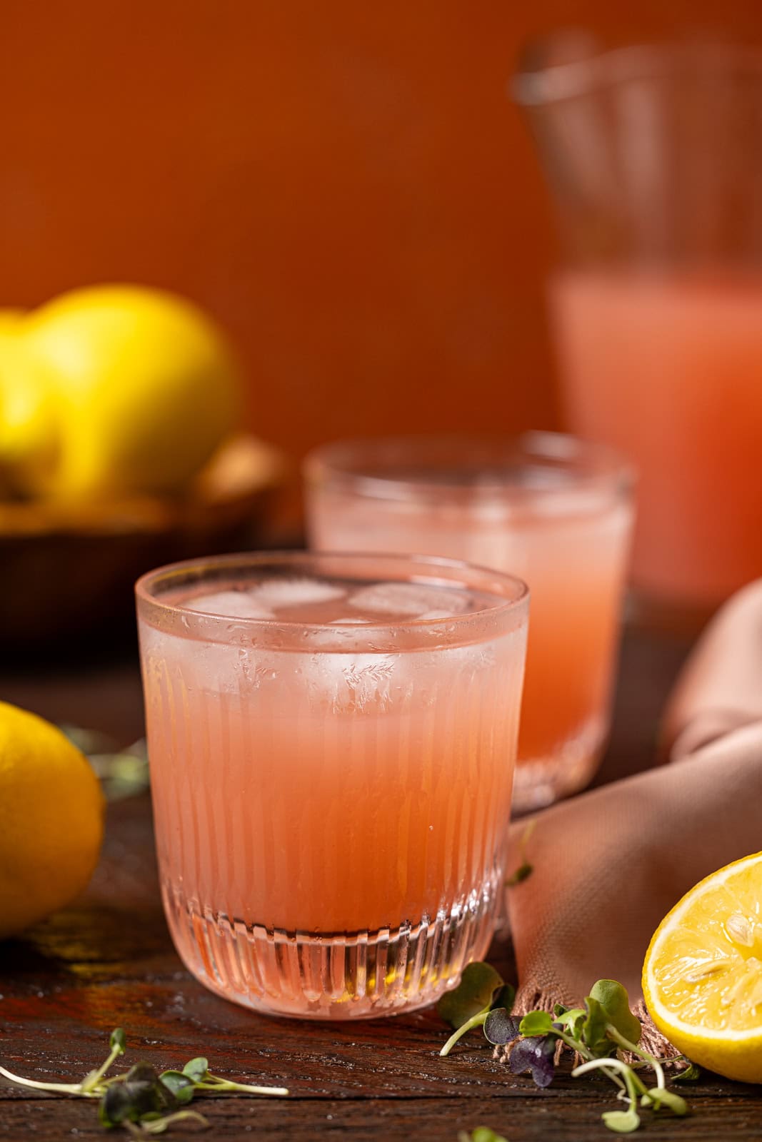 Glass of pink lemonade with a pitcher and lemons.