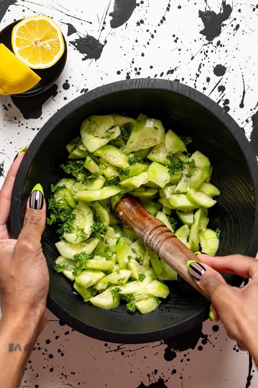 Cucumbers being mashed in a black bowl with a wooden muller.