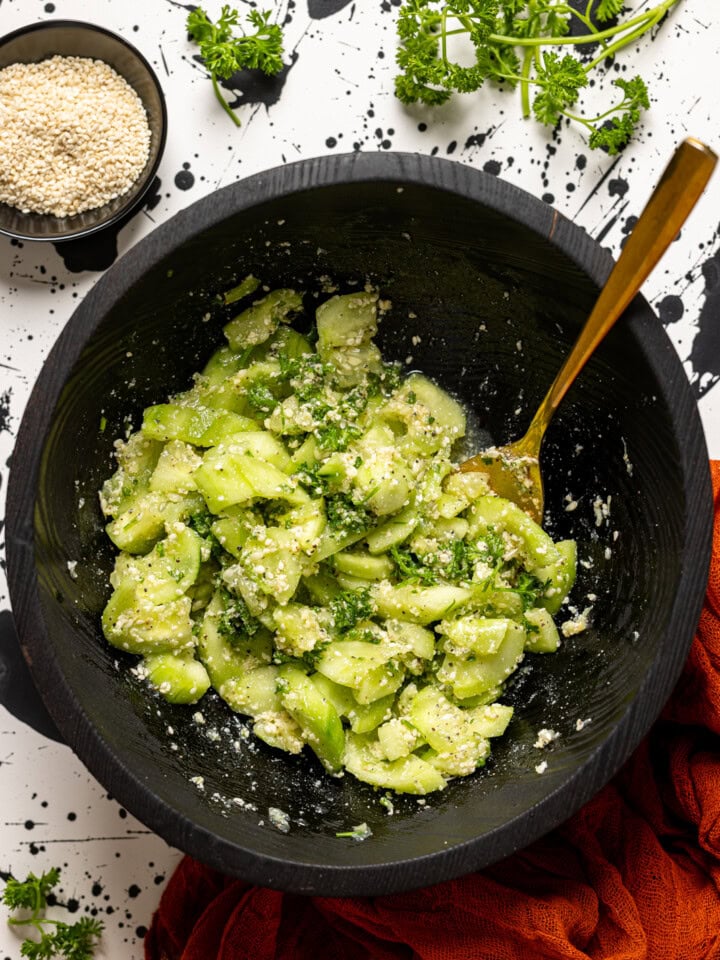 Cucumber salad ingredients in a black bowl with a spoon and side of parmesan.
