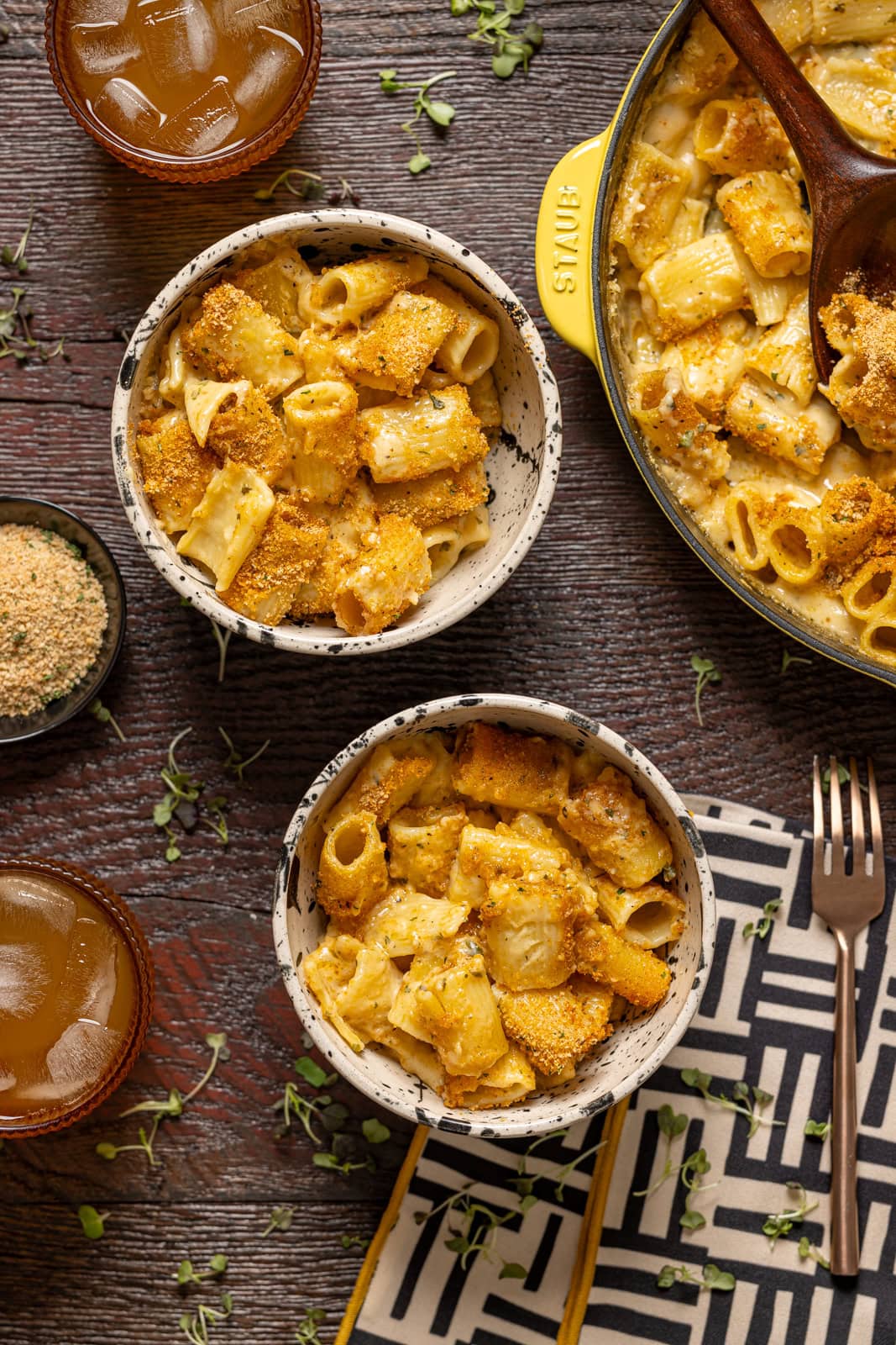 Two bowls of mac and cheese on a brown wood table with a black + white napkin and a side of drink.