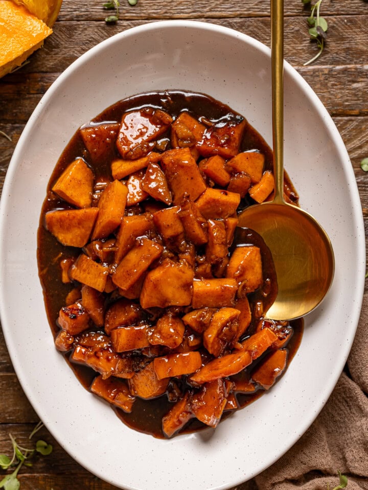 Cooked yams in a white platter with a spoon on a brown wood table.