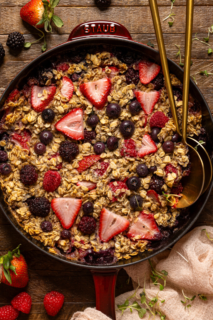 Baked oatmeal in a skillet with gold serving spoons and fresh berries.