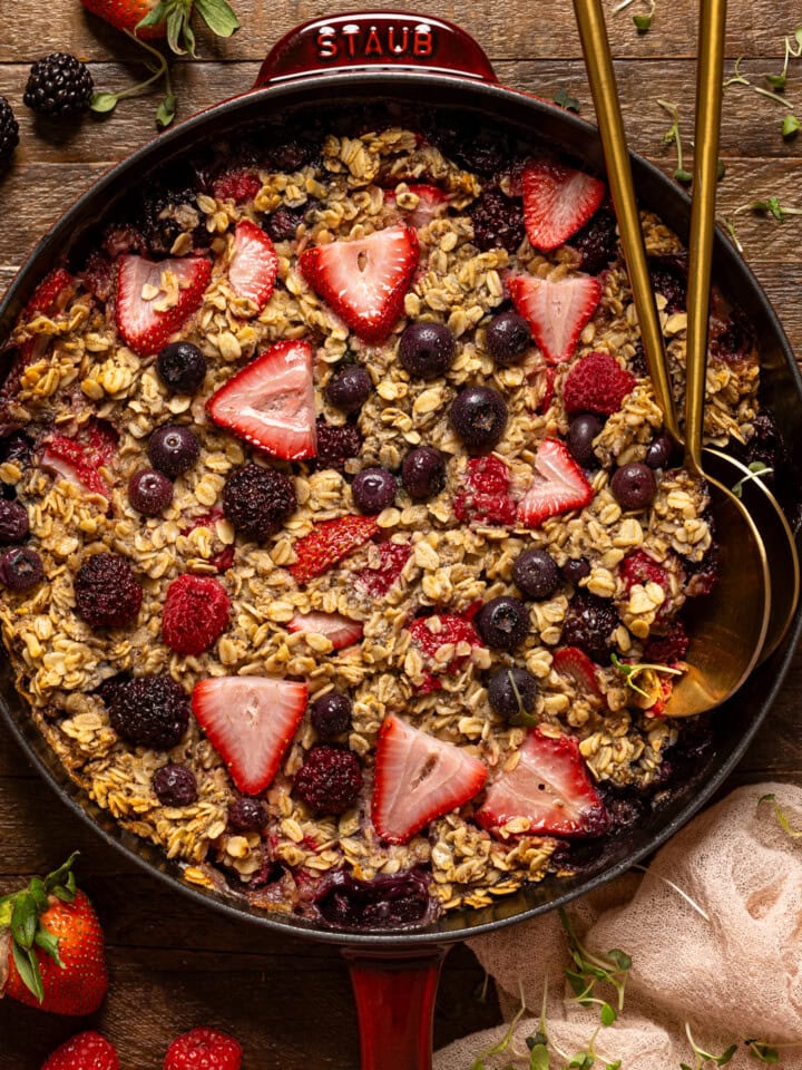 Baked oatmeal in a skillet with gold serving spoons and fresh berries.