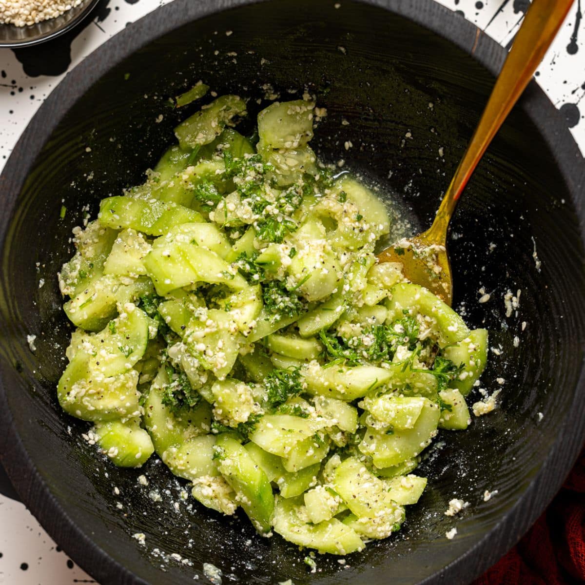 Cucumber salad ingredients in a black bowl with a spoon and side of parmesan.