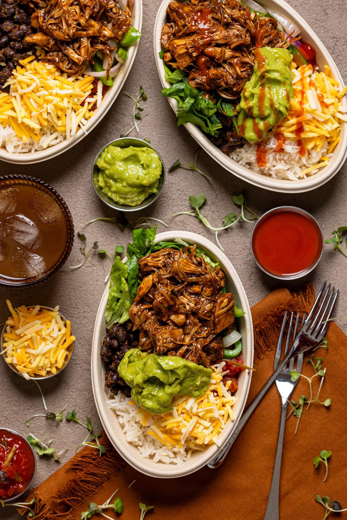 Three burrito bowls with a drink, fork, and condiments.