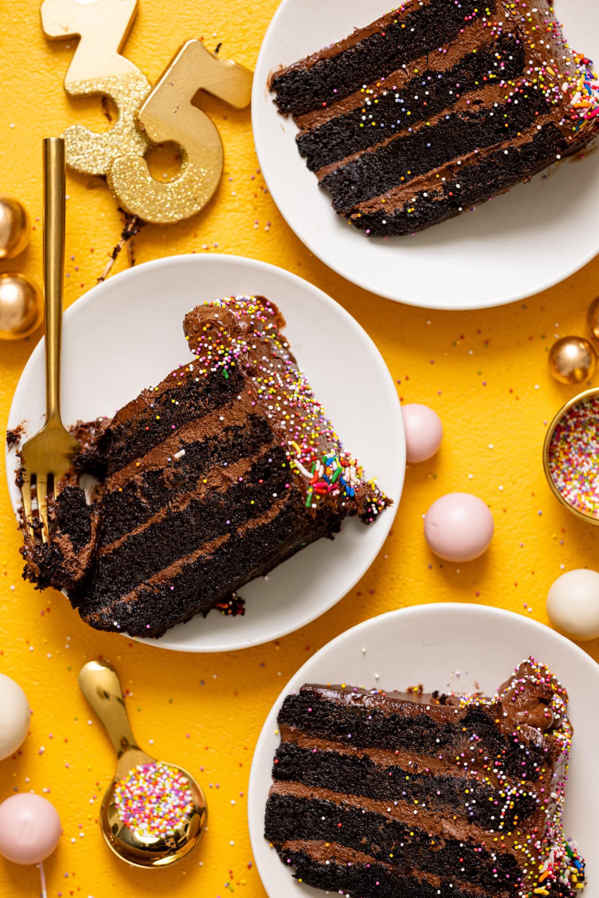 Overhead view of 3 chocolate cake slices on white plates on a yellow table with candles and a fork.