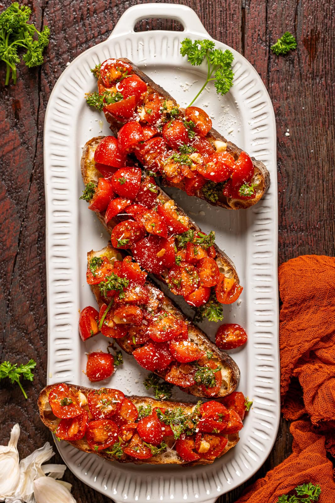 Bruschetta on a platter with fresh parsley on a burgundy wood table.