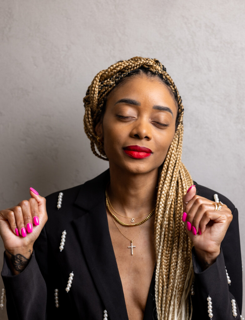 Shanika standing in front of a light grey wall with both hands halfway up with eyes closed in a black + white suit.