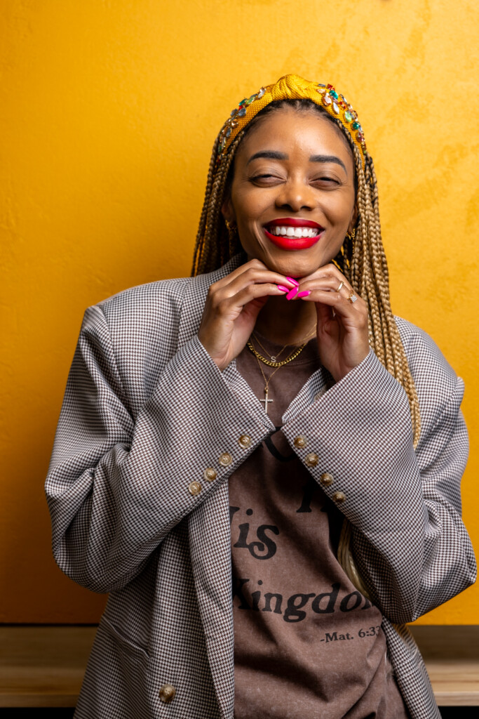 Shanika in front of a yellow wall with a brown shirt and blazer on looking and smiling with hands at chin.