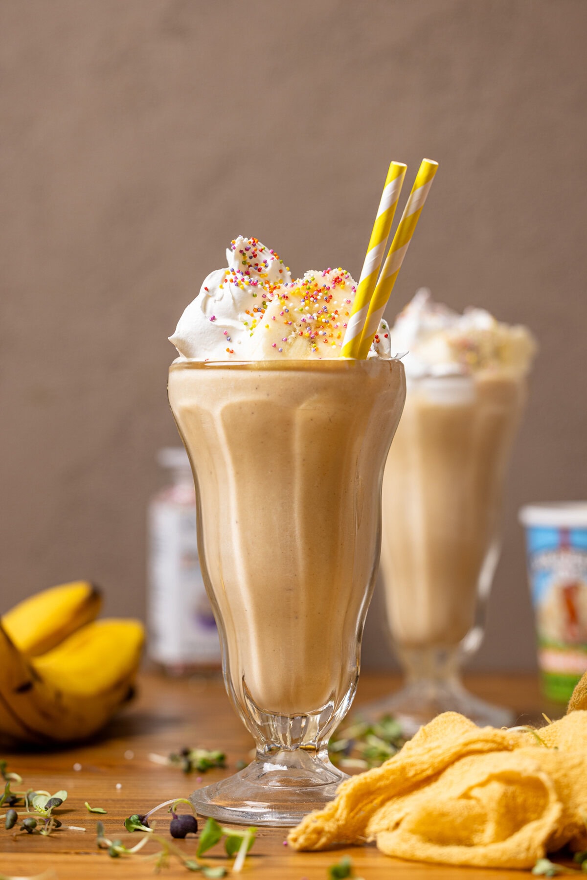 Two glasses filled with milkshake, toppings, bananas, and a yellow napkin.