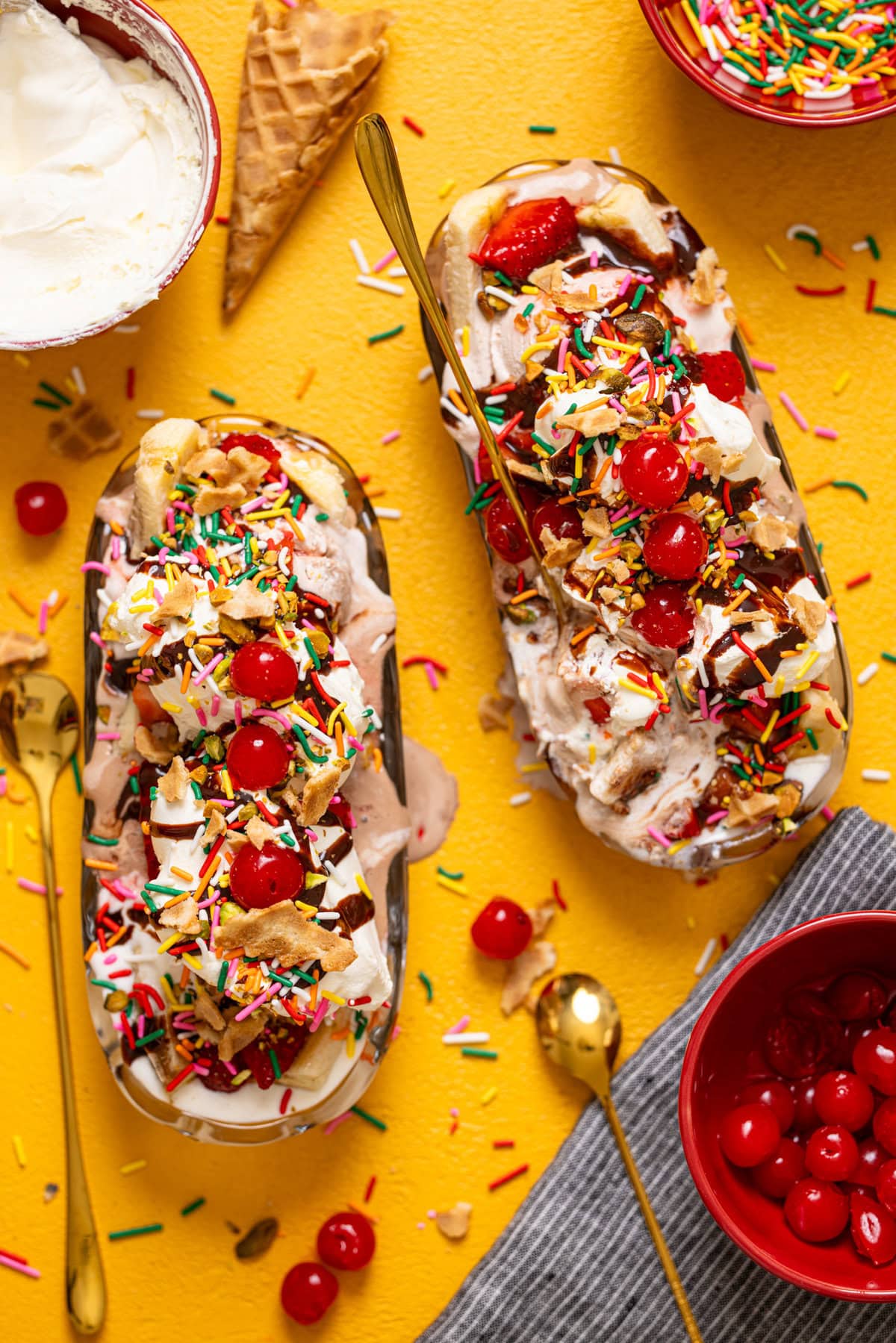 Banana split ice creams on a yellow table with cherries, cones, and more.