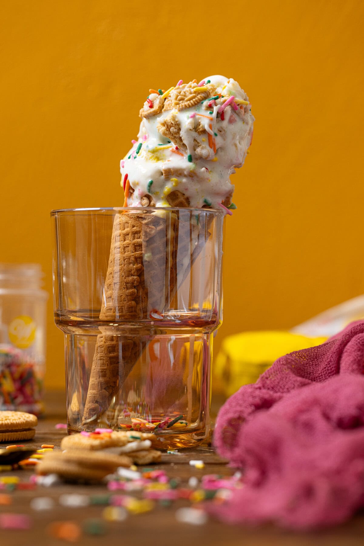 Ice cream cone in glassware in cookies, pink napkin, and sprinkles.
