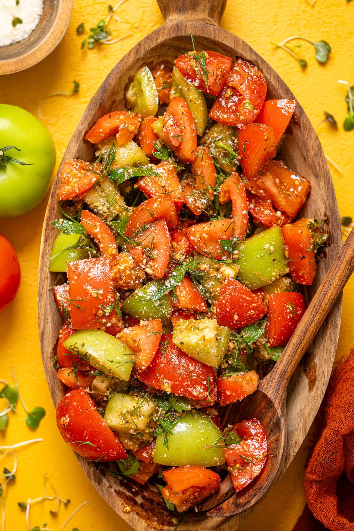 Tomato salad in a wooden serving bowl with a spoon and tomatoes on a yellow table.