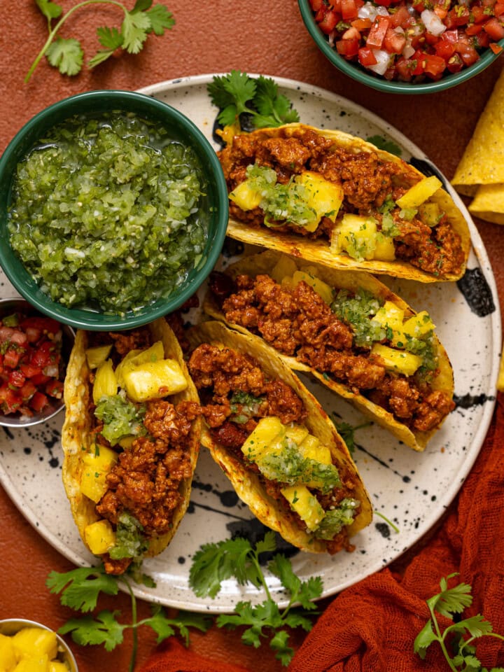 Tacos lined on a plate with verde salsa and sides of toppings.