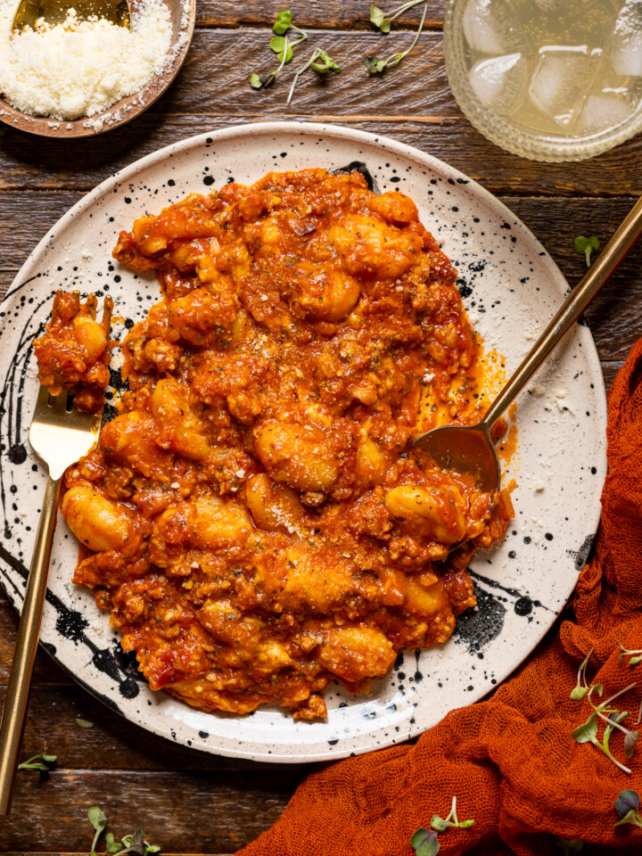An image of baked bolognese gnocchi on a plate with utensils on a wood table with a drink.