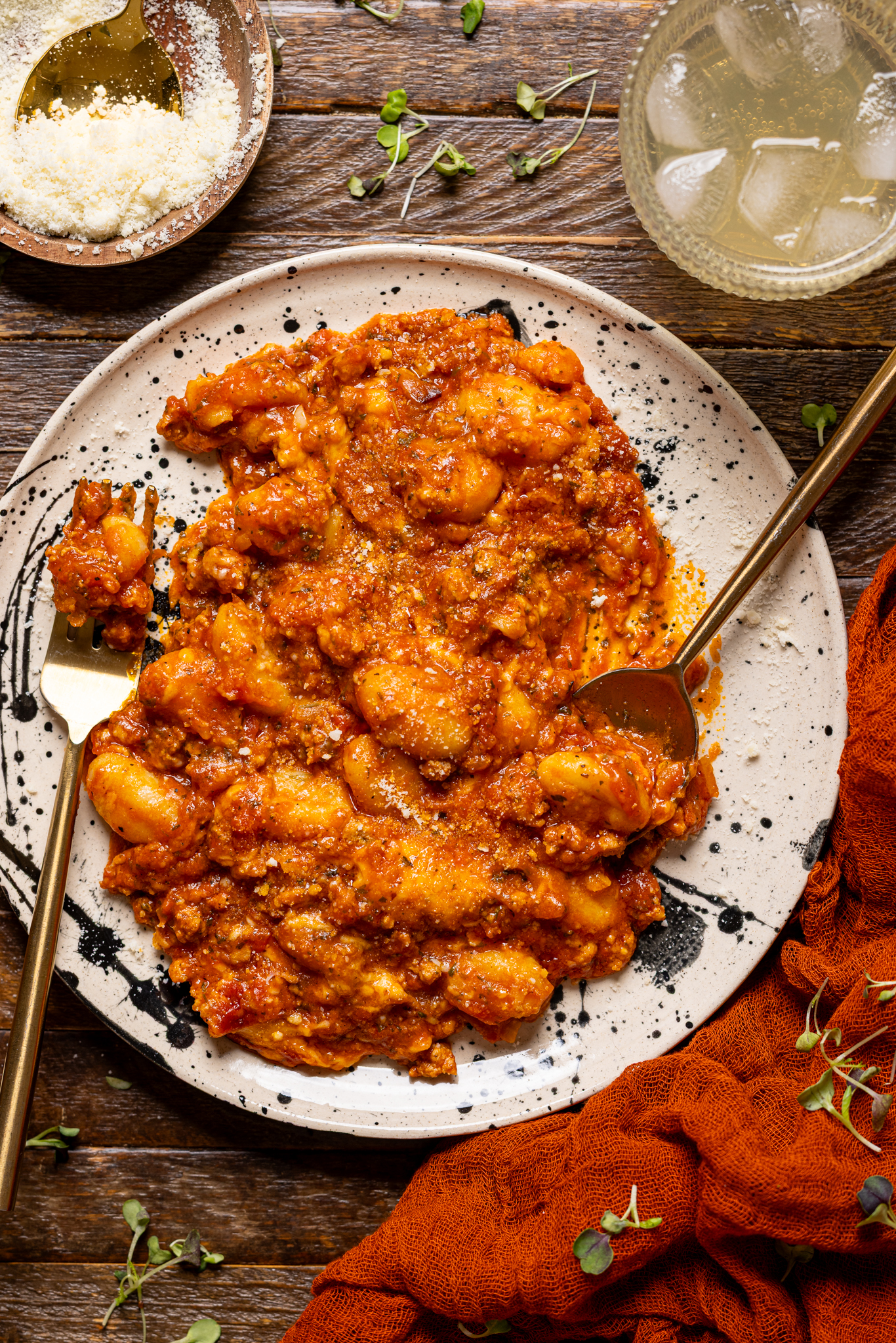 An image of baked bolognese gnocchi on a plate with utensils on a wood table with a drink.