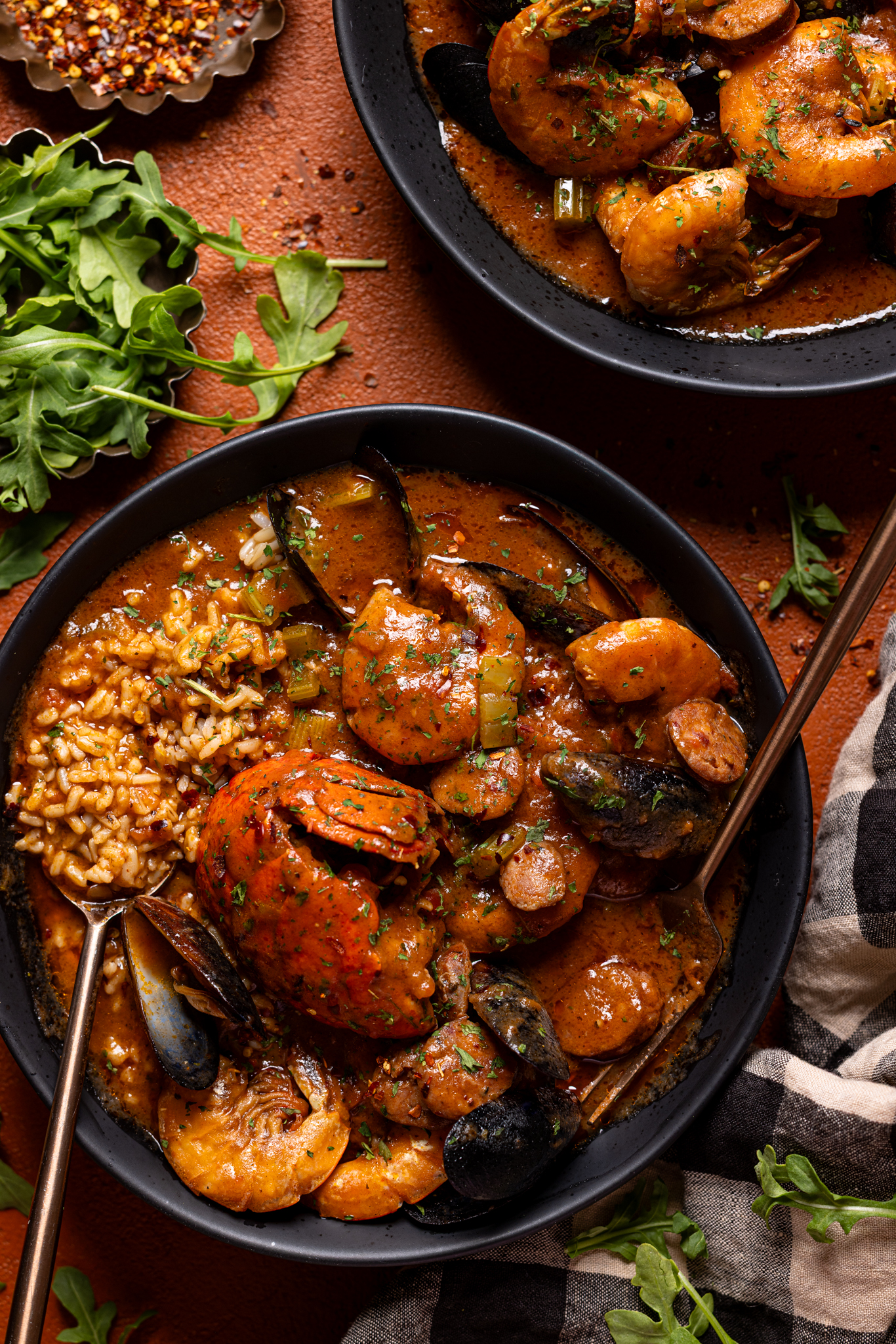 Image of two black bowls filled with seafood gumbo with utensils and herbs.