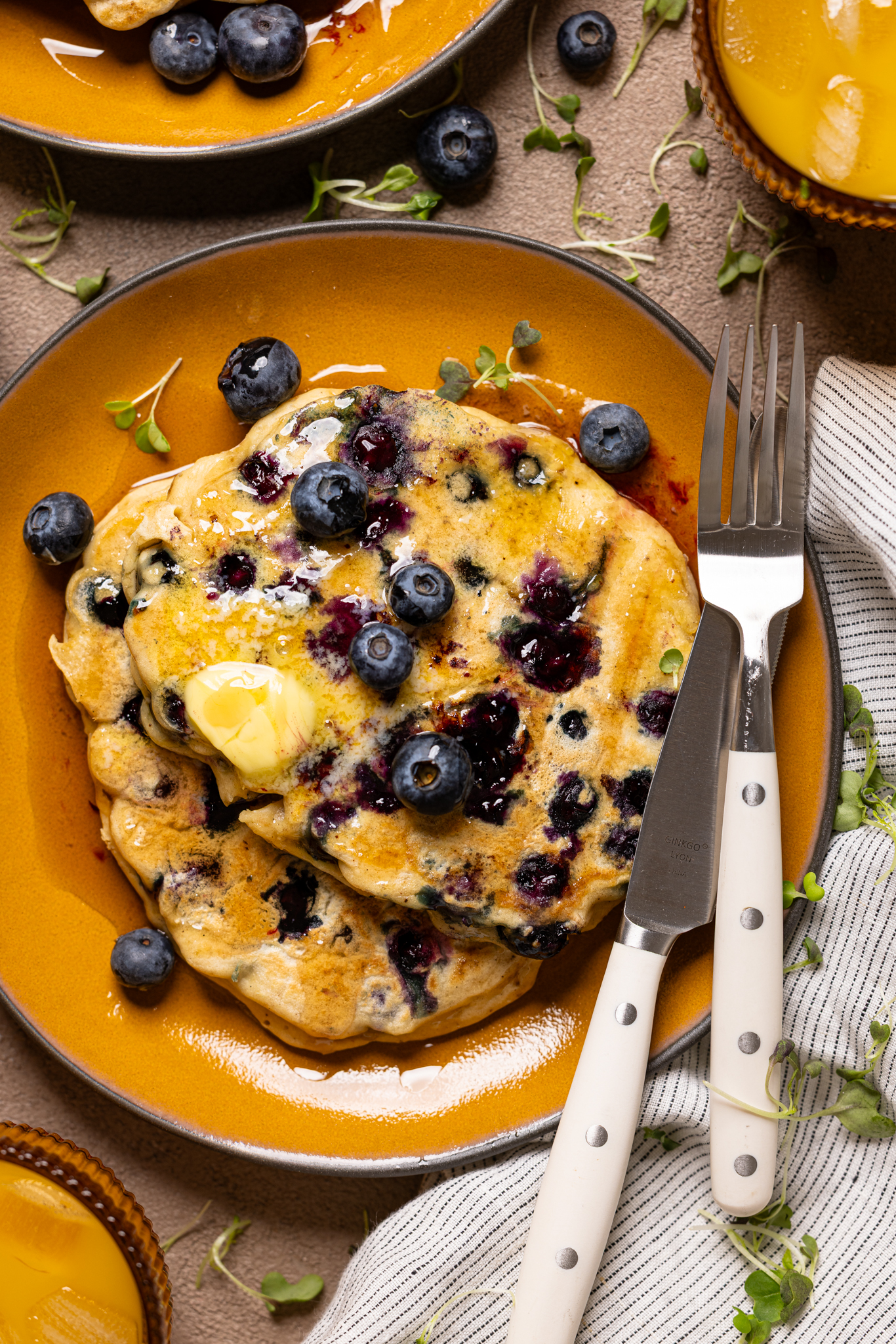 An image of blueberries pancakes on a plate with utensils and orange juice.