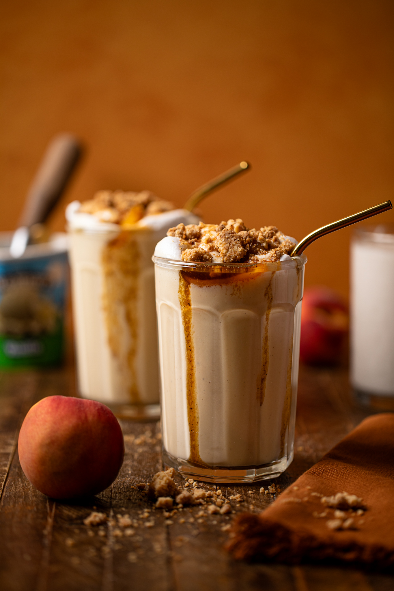 Two glasses of milkshake on a brown wood table with peaches and an ice cream carton.